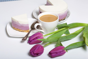 Blueberry cheesecake. A piece of ready-made dessert on a saucer. Jelly layers of different colors are visible. Near a cup of coffee and a bouquet of tulips.
