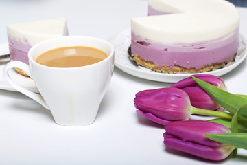 Blueberry cheesecake. A piece of ready-made dessert on a saucer. Jelly layers of different colors are visible. Near a cup of coffee and a bouquet of tulips.