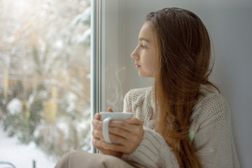 Young woman looking through window enjoys drinking coffee