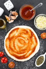 Preparing homemade pizza, ingredients (tomato sauce, garlic, oil, tomato, cheese, oregano, salt, pepper) on the side, photographed overhead on slate