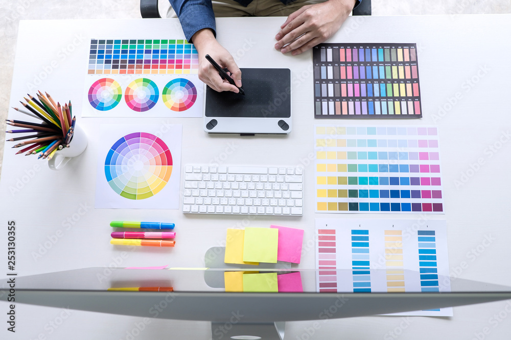 Poster Image of male creative graphic designer working on color selection and drawing on graphics tablet at workplace with work tools and accessories, top view workspace