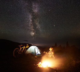 Young woman tourist enjoying at night camping near burning campfire, illuminated tourist tent, mountain bicycle under amazing beautiful evening sky full of stars and Milky way. Astrophotography