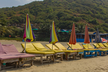 Fototapeta na wymiar multicolored old beach beds with umbrellas on the sand against a green hill with huts