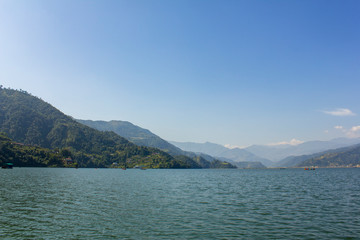 Phewa lake with boats on the background of a green mountain valley under the blue sky, view from the water