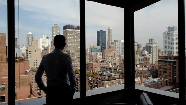 Rear view - a young man stock exchange worker admiring the view of Manhattan and Wall Street from his office with large windows