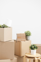 Cardboard boxes of things are stacked on the floor against a white wall. Books and table lamps and green plants in pots. The concept of moving to a new home.