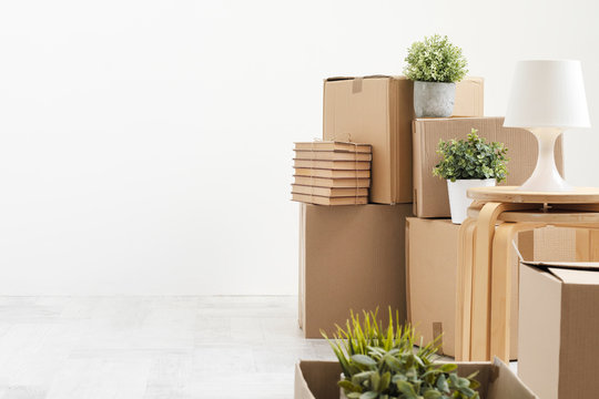 Cardboard boxes with things are stacked on the floor against the background of a white wall close up. Books and table lamps and green plants in pots. The concept of moving to a new home.