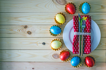 Table setting for Easter - a white plate on it a red napkin with tableware, next to a flower narcissus. A semicircle from the plate are colored eggs of yellow, blue and red. Wooden background.