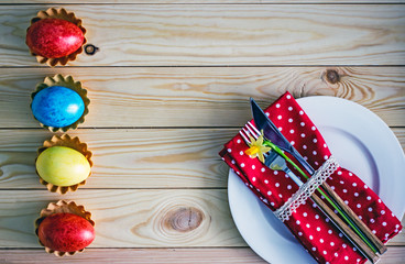 Easter table. А white plate on it a red napkin with tableware, next to a flower narcissus.  Near the plate are colored eggs of yellow, blue and red. Easter card. View from above.