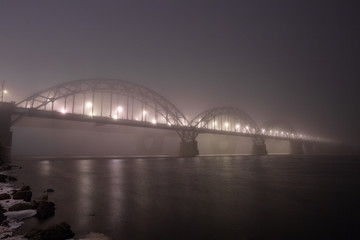 Fototapeta na wymiar A mysterious evening fog above the river in big city. Bridge in the mist, cold weather scenery. Soft, blurry, misty look. Colorful, mystic industrial cityscape.