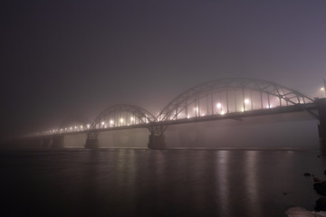 Naklejka premium A mysterious evening fog above the river in big city. Bridge in the mist, cold weather scenery. Soft, blurry, misty look. Colorful, mystic industrial cityscape.