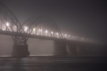 Obraz premium A mysterious evening fog above the river in big city. Bridge in the mist, cold weather scenery. Soft, blurry, misty look. Colorful, mystic industrial cityscape.
