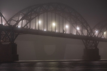 A mysterious evening fog above the river in big city. Bridge in the mist, cold weather scenery. Soft, blurry, misty look. Colorful, mystic industrial cityscape.