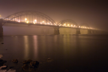 Fototapeta premium A mysterious evening fog above the river in big city. Bridge in the mist, cold weather scenery. Soft, blurry, misty look. Colorful, mystic industrial cityscape.