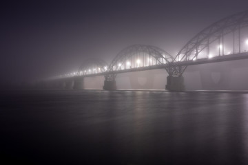 Obraz premium A mysterious evening fog above the river in big city. Bridge in the mist, cold weather scenery. Soft, blurry, misty look. Colorful, mystic industrial cityscape.