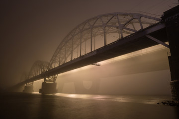 Obraz premium A mysterious evening fog above the river in big city. Bridge in the mist, cold weather scenery. Soft, blurry, misty look. Colorful, mystic industrial cityscape.