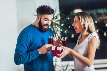 Beautiful young couple is celebrating at home. Pretty girl is giving her boyfriend a gift box