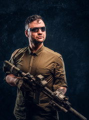 Fototapeta na wymiar Portrait of a stylish man wearing shirt sunglasses holding assault rifle and looking sideways. Studio photo against a dark textured wall