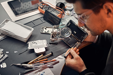 Serviceman uses magnifier and screwdriver to repair damaged smartphone in the electronic workshop.