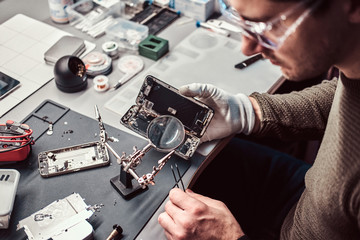 Serviceman uses magnifier and tweezers to repair damaged smartphone in the electronic workshop.