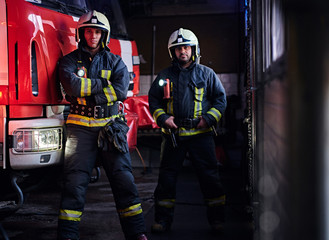 Two firemen wearing protective uniform standing next to a fire engine in a garage of a fire...