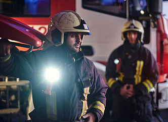 Two brave firemen wearing a protective uniform standing next to a fire truck. Arrival on call at...