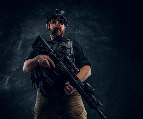 Fototapeta na wymiar Bearded special forces soldier or private military contractor holding an assault rifle and observes the surroundings in night vision goggles. Studio photo against a dark textured wall