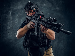 Fototapeta na wymiar Bearded special forces soldier wearing body armor and helmet with night vision holding an assault rifle. Studio photo against a dark textured wall