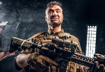 Fototapeta na wymiar Crazy special forces soldier wearing body armor holding assault rifle. Studio photo against a dark textured wall