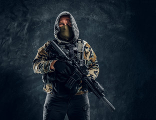 Special forces soldier in military uniform wearing mask and hood holding an assault rifle. Studio photo against a dark textured wall