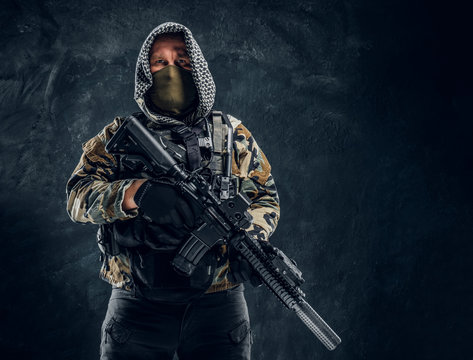 Special forces soldier in military uniform wearing mask and hood holding an assault rifle. Studio photo against a dark textured wall