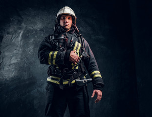 Firefighter in full protective equipment holding an oxygen mask and looking at a camera. Studio photo against a dark textured wall