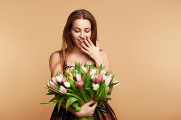 Wow effect. Wonderful spring flowers on Womens Day. Tender surprised brunet woman poses with colorful tulips, looks happily, isolated over beige studio background wall. Space for text