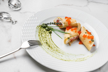 Crepes with filet salmon and cheese, red fish caviar and green sauce on white plate over marble background. Pancake week or Shrovetide. Healthy breakfast