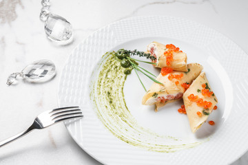 Crepes with filet salmon and cheese, red fish caviar and green sauce on white plate over marble background. Pancake week or Shrovetide. Healthy breakfast. Top view, flat lay
