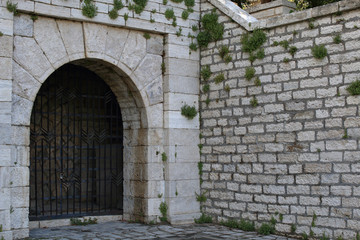 Entrance arch with metal bars and old white stone wall with plants between bricks