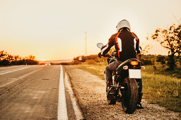 Full length portrait from the back of a european biker on his motorcycle being ready to start his traveling with his bike to find new places against the sunset.