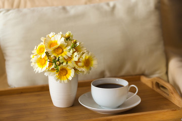 Obraz na płótnie Canvas A cup of tea / coffee and a bouquet of daisies in a vase on a wooden tray against the background of a light soft pillow