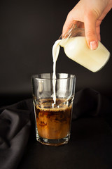 Iced coffee being poured in a tall glass with ice. Pouring milk in glass of coffee. Cold, refreshing, caffeine milkshake. Summmer drink. Black background.