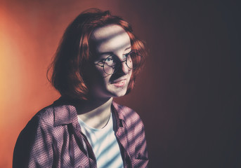 Red-haired girl with glasses in a plaid shirt and a white t-shirt is sad at the window with blinds