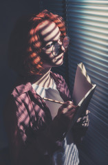 A red-haired girl in a plaid shirt and a white t-shirt draws in a notebook with a pencil, standing...