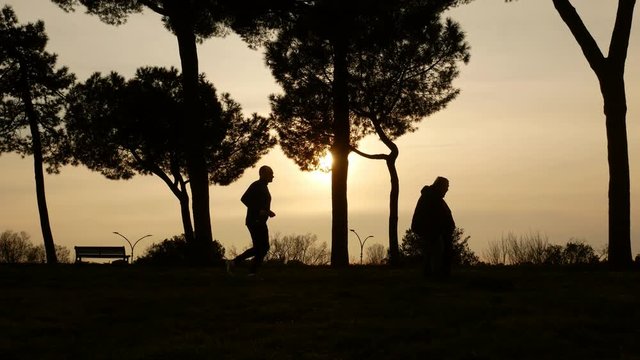 A man jog and a couple walk in a park, high contrast backlight silhouette at sunset