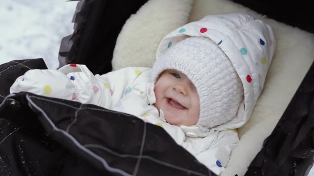 A close-up plan of a laughing baby in the warm hat sitting in a black pram in a winter park