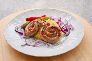 Obraz na płótnie Canvas Czech traditional and seasonal spiral fried fresh white wine sausages with mashed potatoes, pickles from cucumber and hot pepper, chopped fresh red onion served on white plate