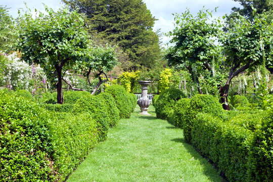 Stone flowerpot in the middle of an English garden in bloom at the end of grass footpath between trimmed topiary green plants .