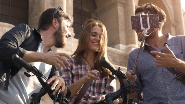Three happy young friends tourists with bikes and backpacks at Colosseum in Rome taking selfies pictures with smartphone and stick having fun on sunny day slow motion steadycam