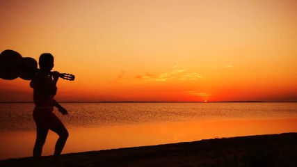 Young girl walking with guitar on the beach at sunset