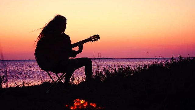 Young girl playing guitar on the beach with campfire at sunset
