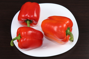 Three large red peppers lie on a white serving plate, standing on a table with a dark background.