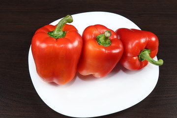 Three large red peppers lie on a white serving plate, standing on a table with a dark background.